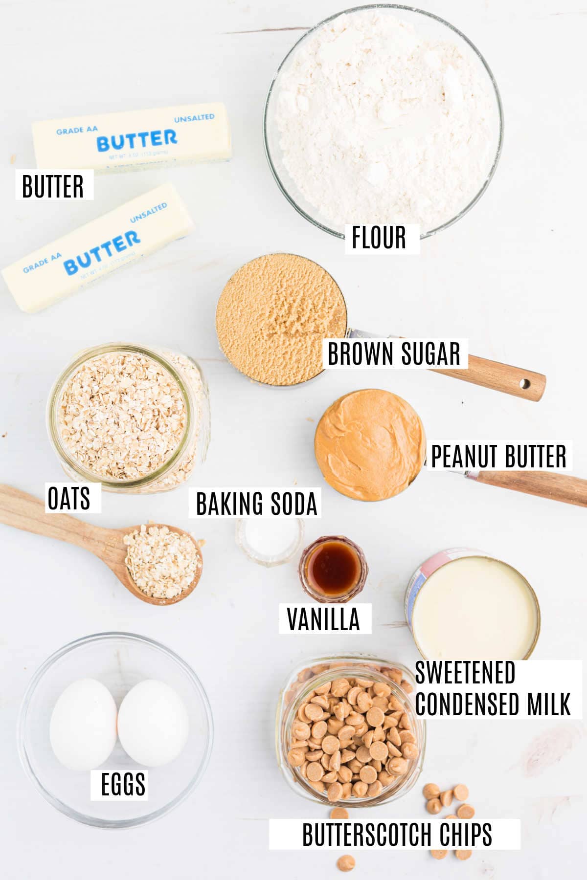 Baking ingredients on a white surface—perfect for making peanut butter revel bars—including butter, flour, brown sugar, oats, peanut butter, eggs, and more, all labeled.