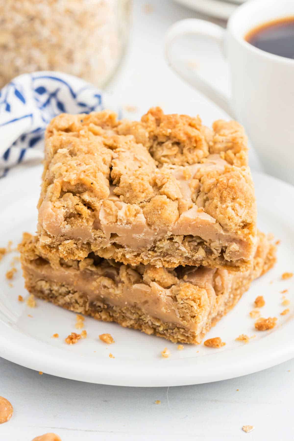 Two oatmeal caramel bars stacked on a white plate next to a cup of coffee and a blue-white napkin, reminiscent of classic peanut butter revel bars.