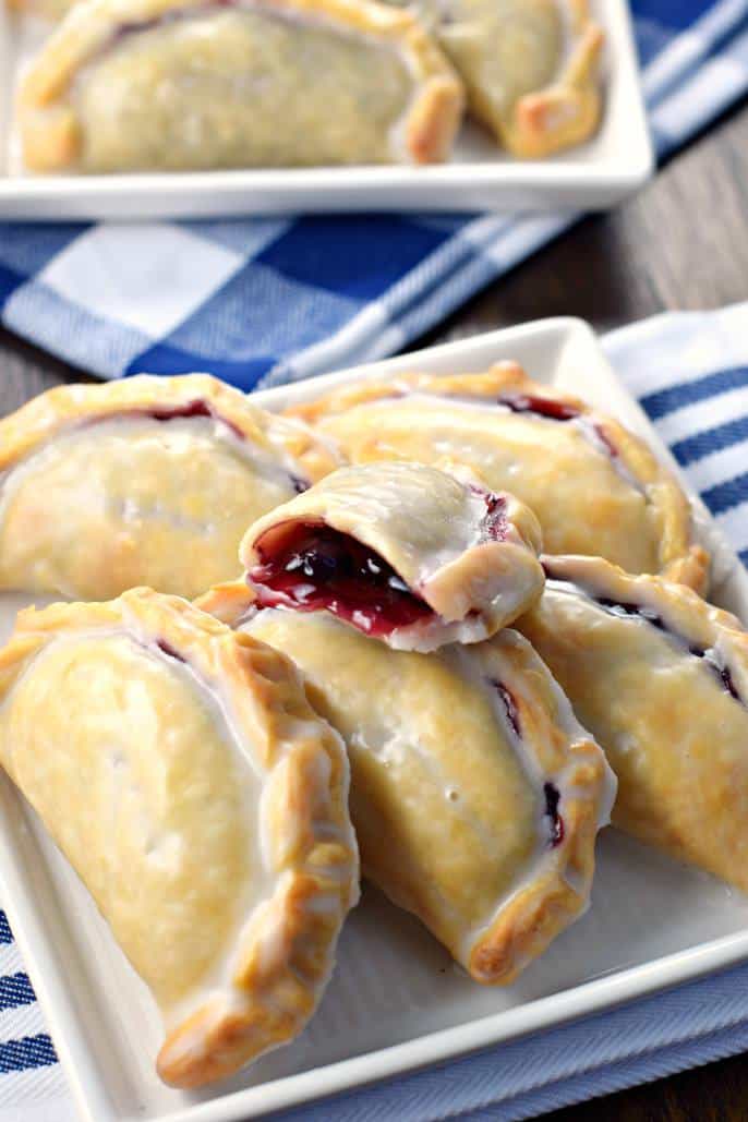 Blueberry hand pies on a white plate. One hand pie cut in half with pie filling oozing out.