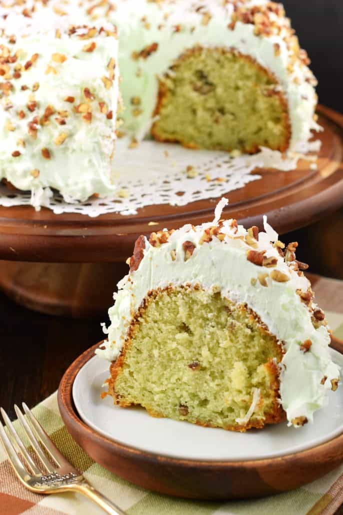 Slice of pistachio cake with pistachio frosting on a white plate with whole cake in background.