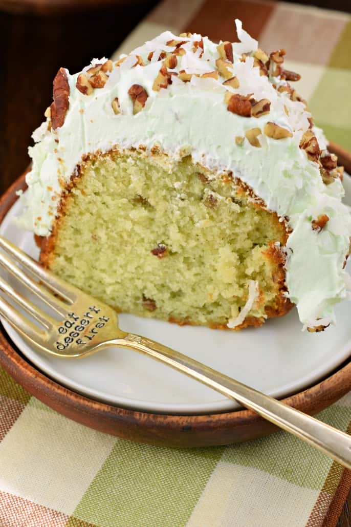 Slice of pistachio cake with pistachio frosting on a white plate and fork that says "eat dessert first."