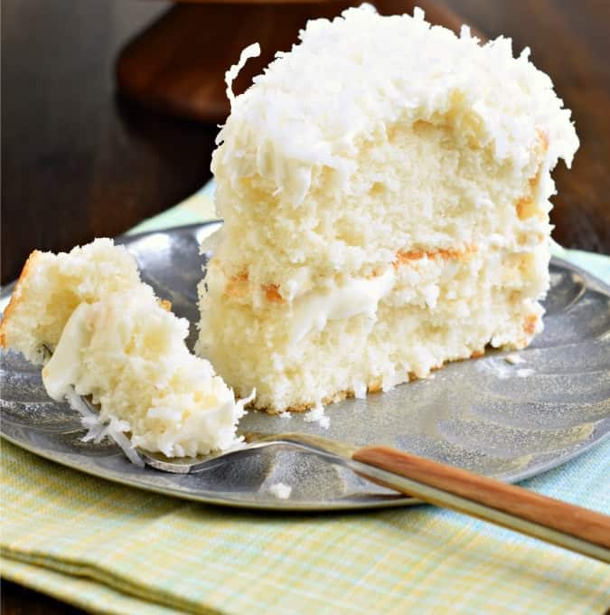 Slice of coconut cake on a silver plate with one forkful taken away.