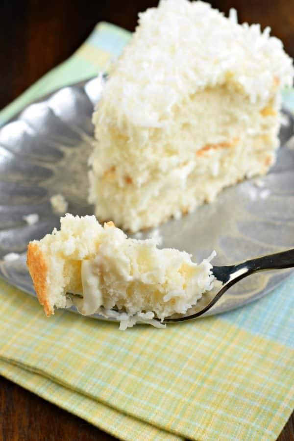 Forkful of coconut cake with a slice on a silver plate.