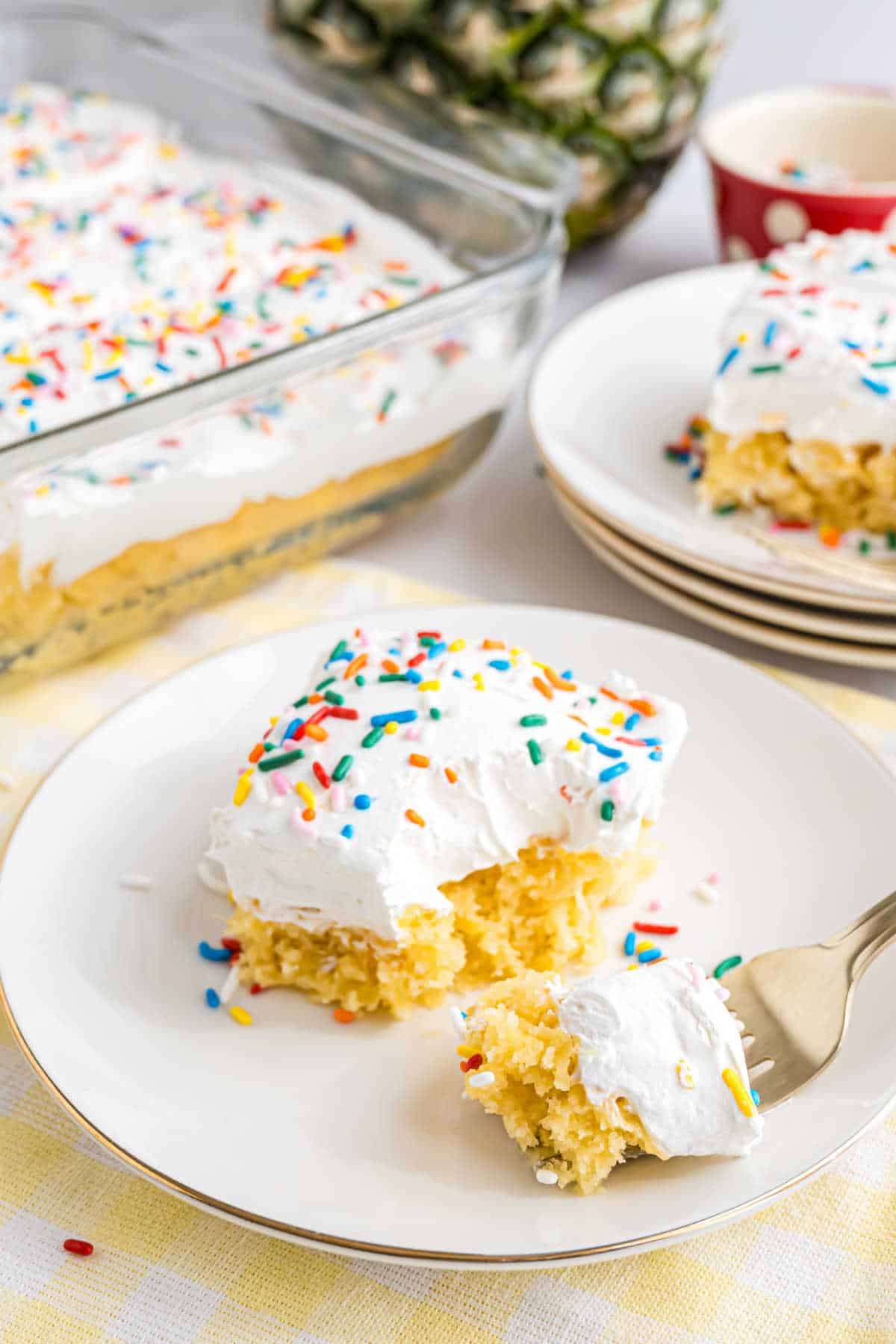 A slice of skinny pineapple cake with white frosting and rainbow sprinkles sits on a plate, with a fork and more cake in the background.