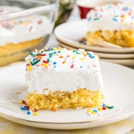 A slice of skinny pineapple cake with white frosting and colorful sprinkles rests on a white plate.