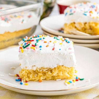 A slice of skinny pineapple cake with white frosting and colorful sprinkles rests on a white plate.