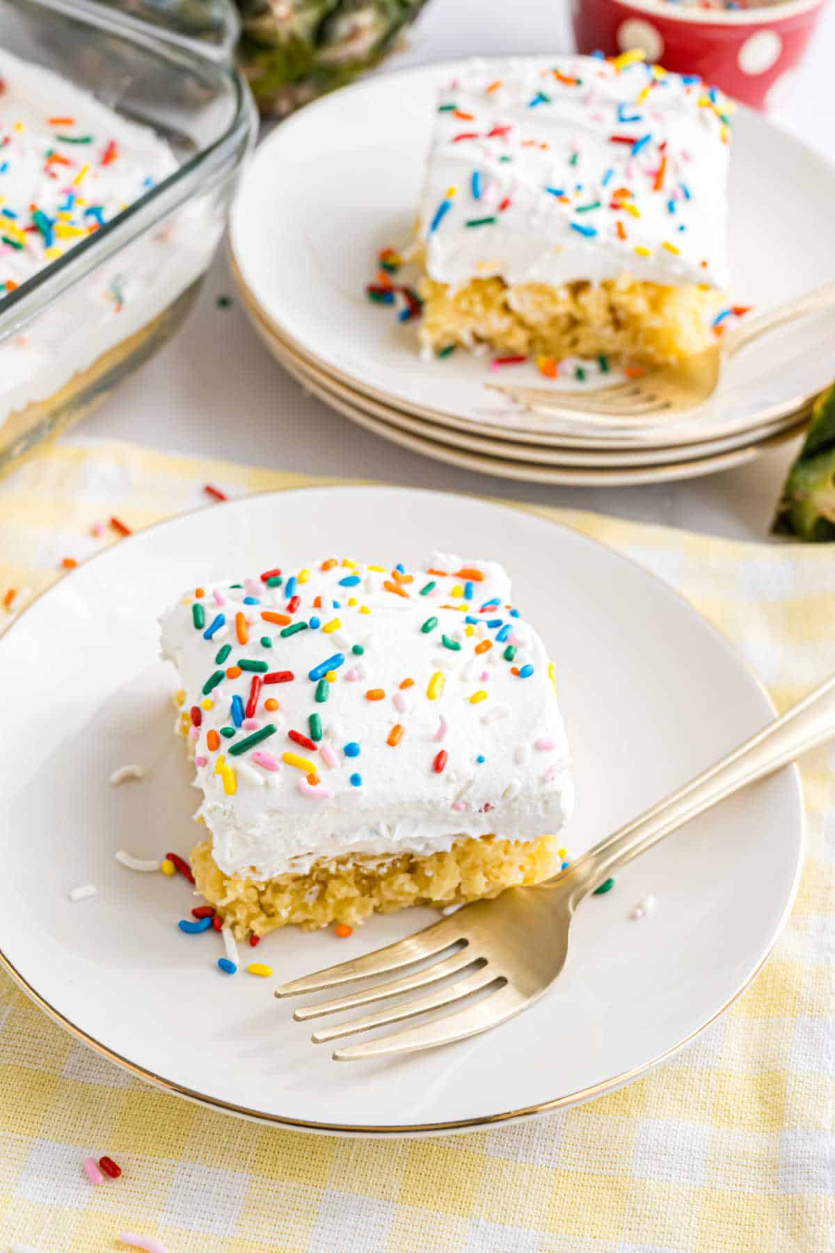 A slice of skinny pineapple cake with white frosting and colorful sprinkles sits on a white plate with a gold fork.