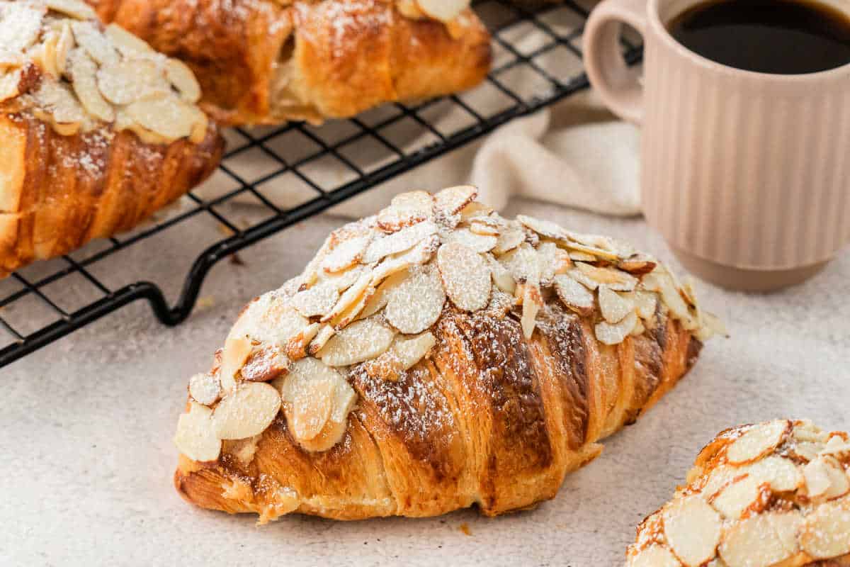 Easy almond croissants topped with sliced almonds and powdered sugar, next to a mug of coffee on a light surface.