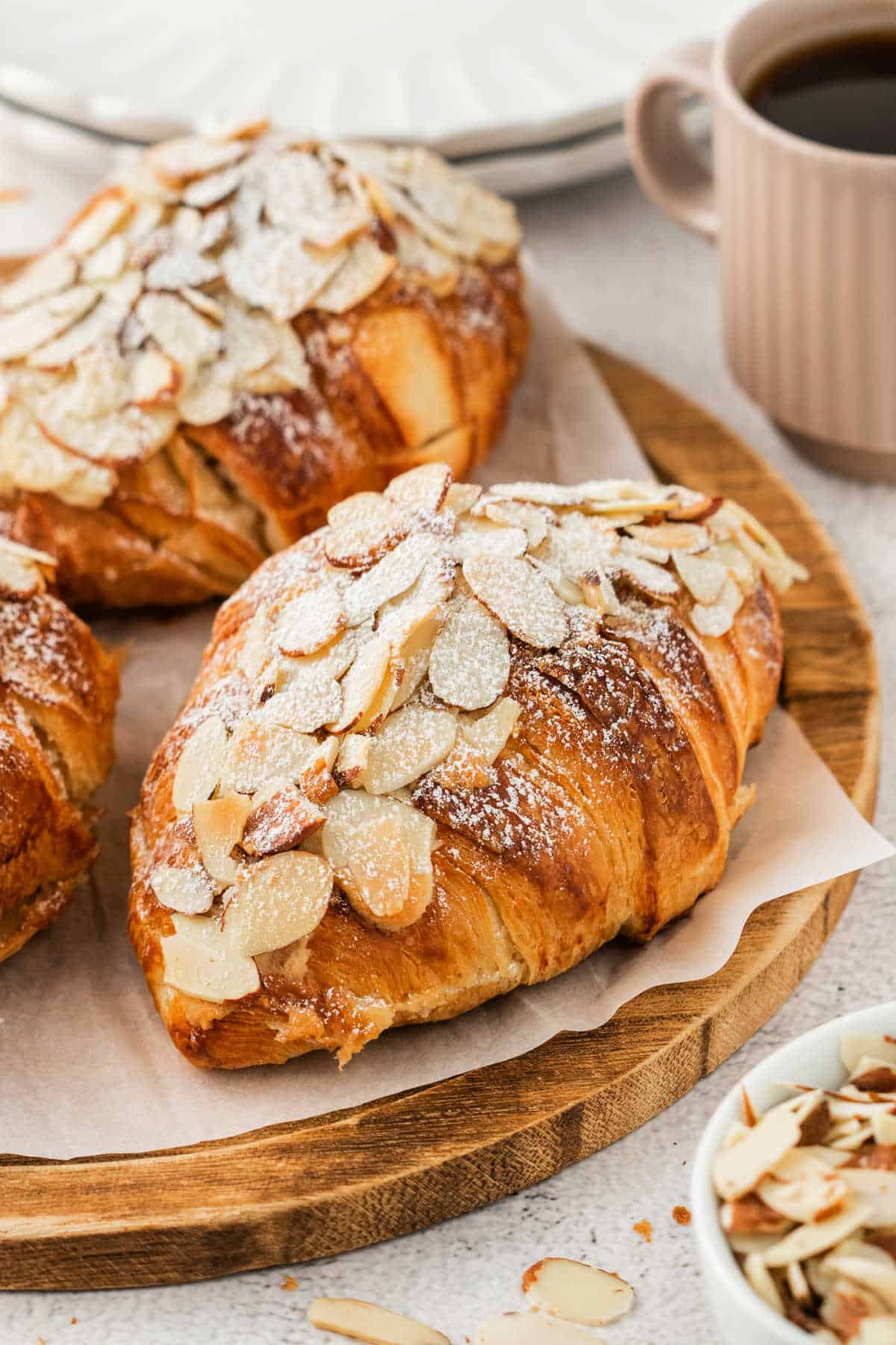 Enjoy easy almond croissants topped with sliced almonds and powdered sugar, served on parchment beside a cup of coffee and extra sliced almonds.