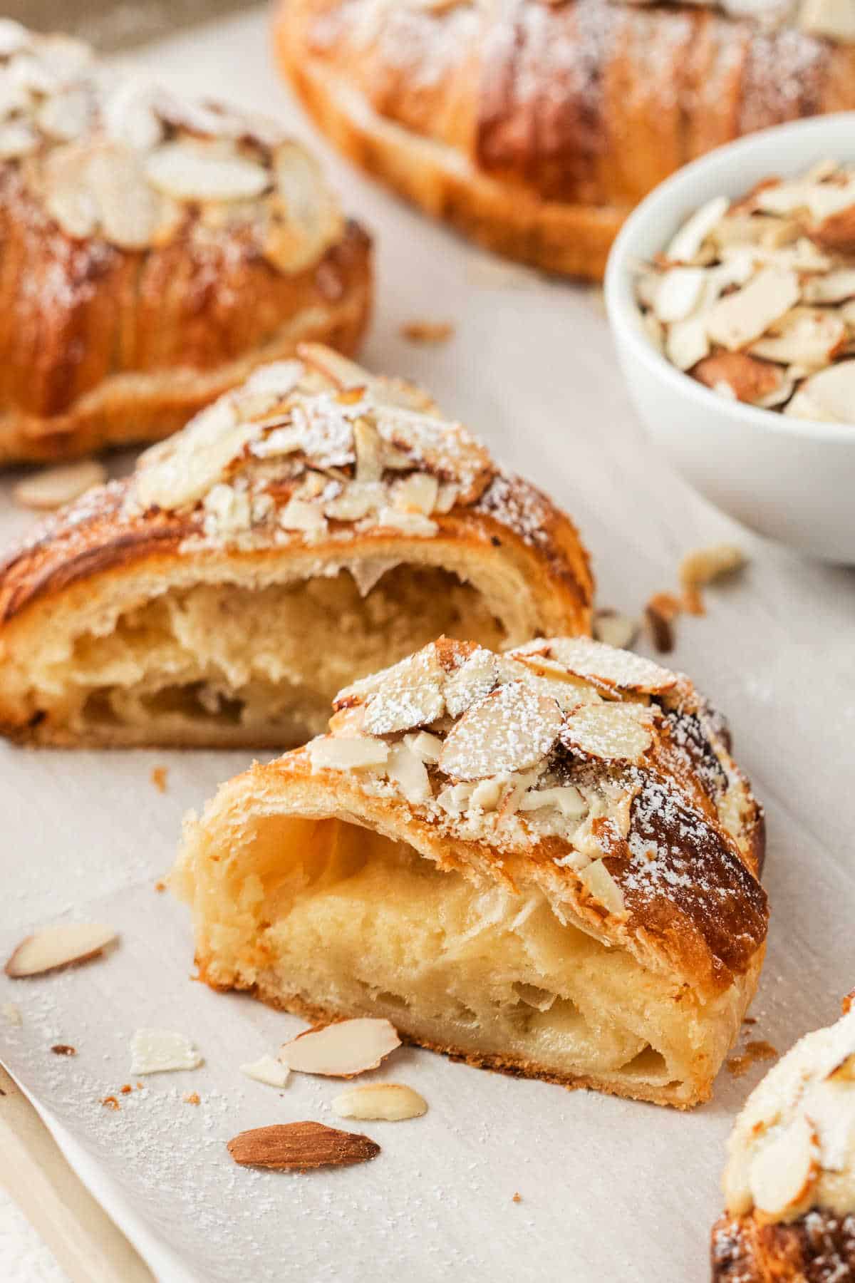 An easy almond croissant, sliced and topped with powdered sugar and almond flakes, sits on parchment paper.