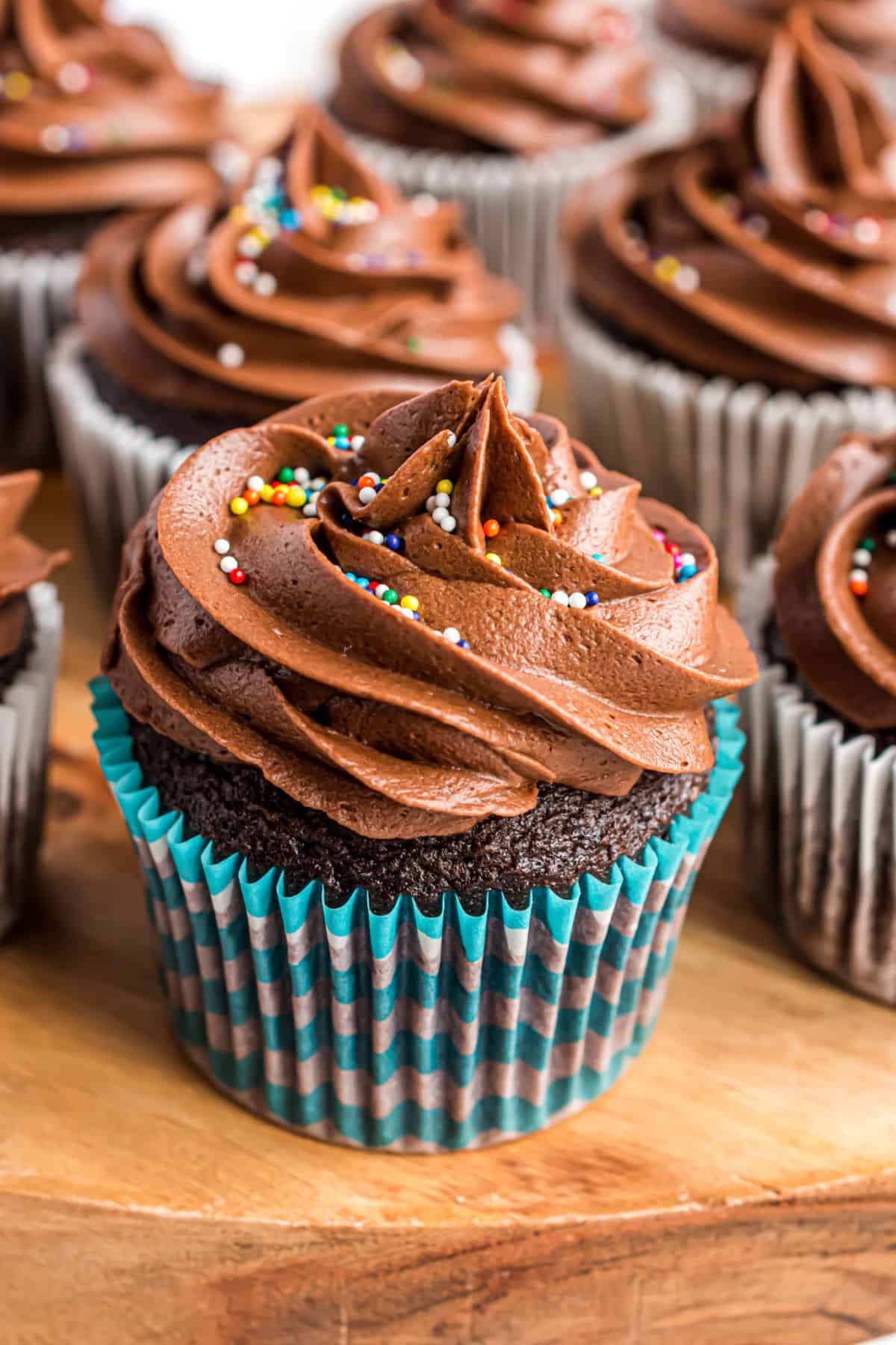 Chocolate frosted chocolate cupcakes with colorful sprinkles on wooden platter.