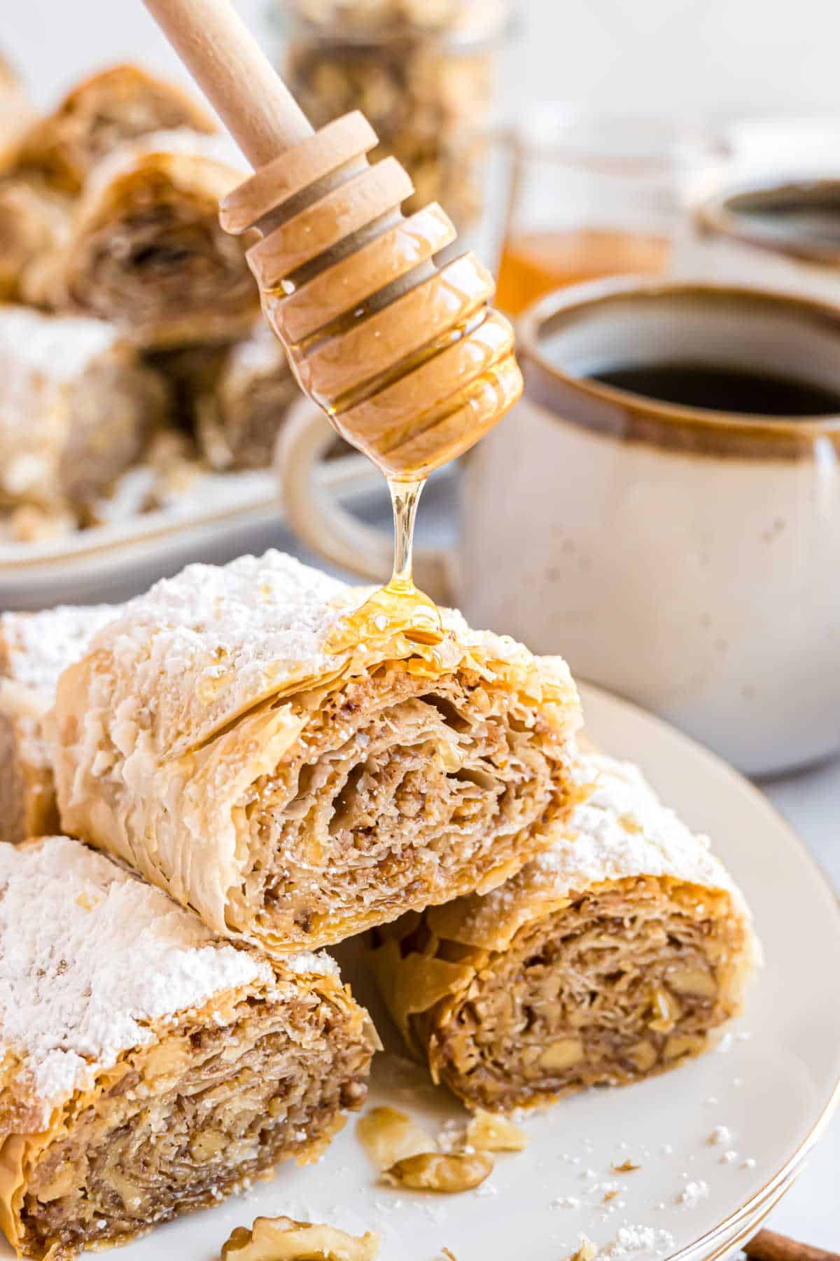 A honey dipper drizzles honey over slices of baklava, the nut-filled pastry dusted with powdered sugar, next to a coffee mug.