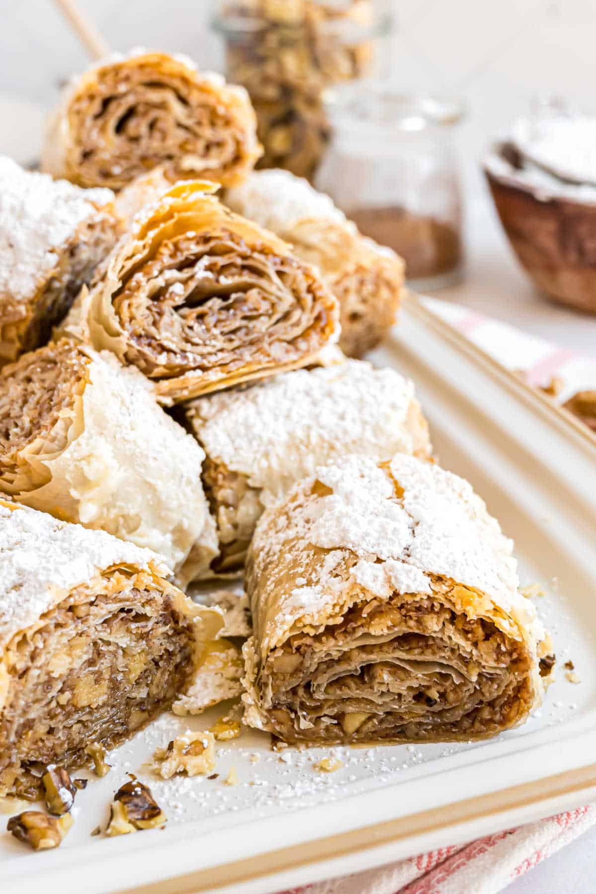Rolled baklava pieces dusted with powdered sugar rest on a tray, with walnuts and jars in the background.
