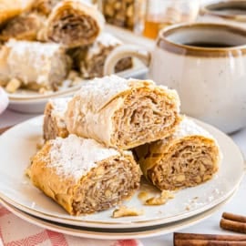 Four slices of baklava topped with powdered sugar rest on a white plate beside a steaming cup of coffee.