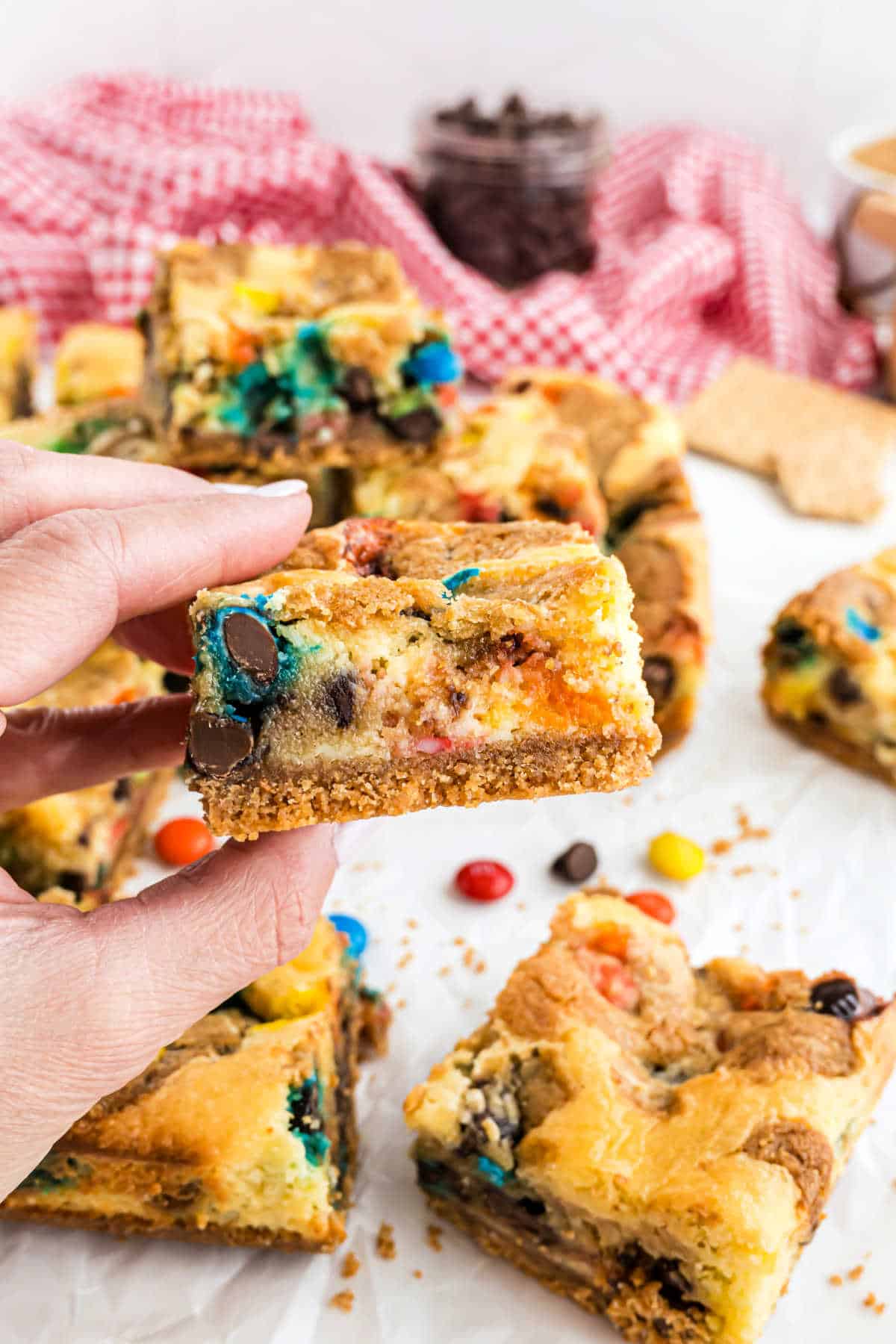 A hand holds a colorful M&M Cheesecake Bar studded with chocolate chips and candy-coated pieces; more cheesecake bars are visible in the background.