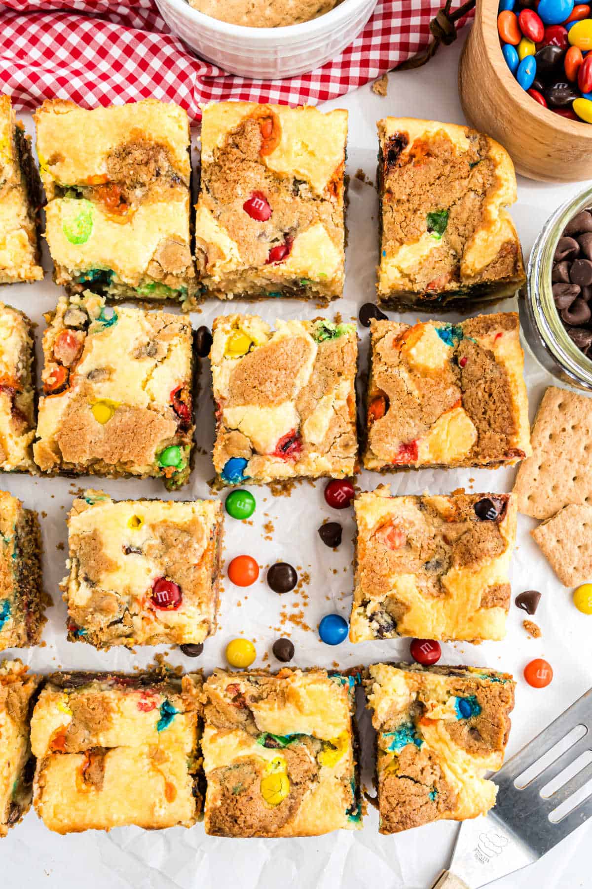 Top view of M&M Cheesecake Bars with chocolate chips, sliced into squares, on a white surface with snacks nearby.