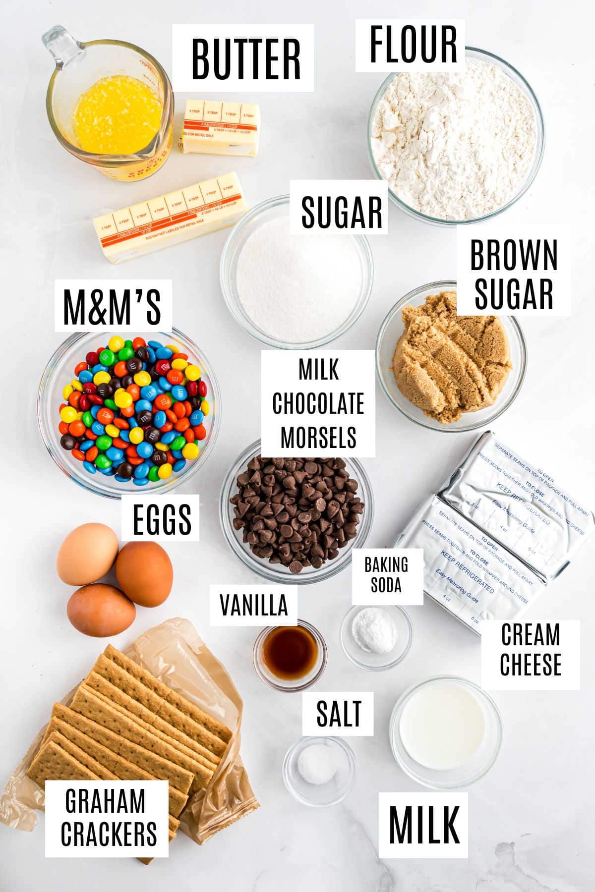 Baking ingredients labeled: butter, flour, sugar, brown sugar, M&M’s, chocolate chips, eggs, vanilla, cream cheese, and milk—everything you need to whip up delicious M&M Cheesecake Bars.