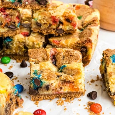 A stack of colorful M&M Cheesecake Bars and chocolate chip cookie bars on parchment paper, with some loose candies scattered around.