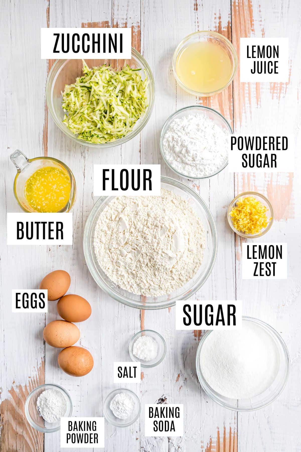 Baking ingredients on a white table, each labeled for making lemon zucchini bread: zucchini, flour, sugar, eggs, butter, lemon juice, and more.