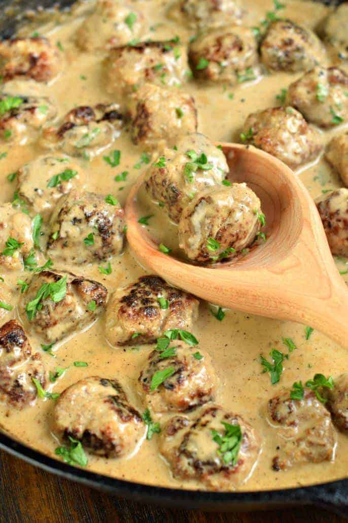 Swedish meatballs in creamy sauce in a black skillet with a wooden spoon. Meatballs garnished with parsley.