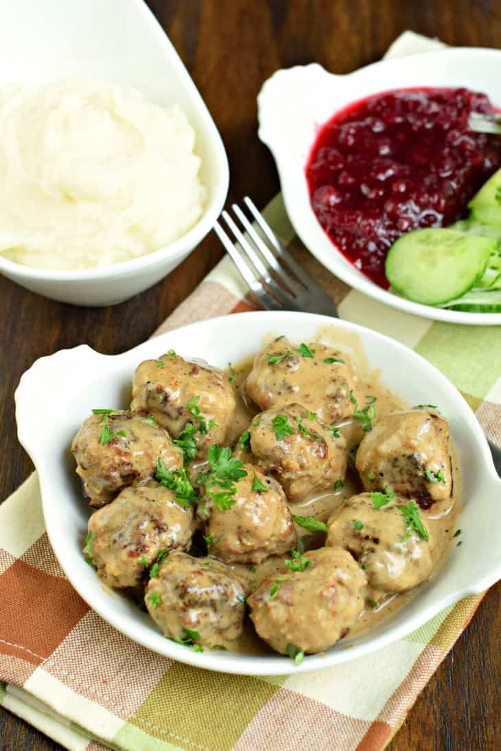 Swedish meatballs in white bowl served with mashed potatoes, cucumber dill salad, and lingonberries.
