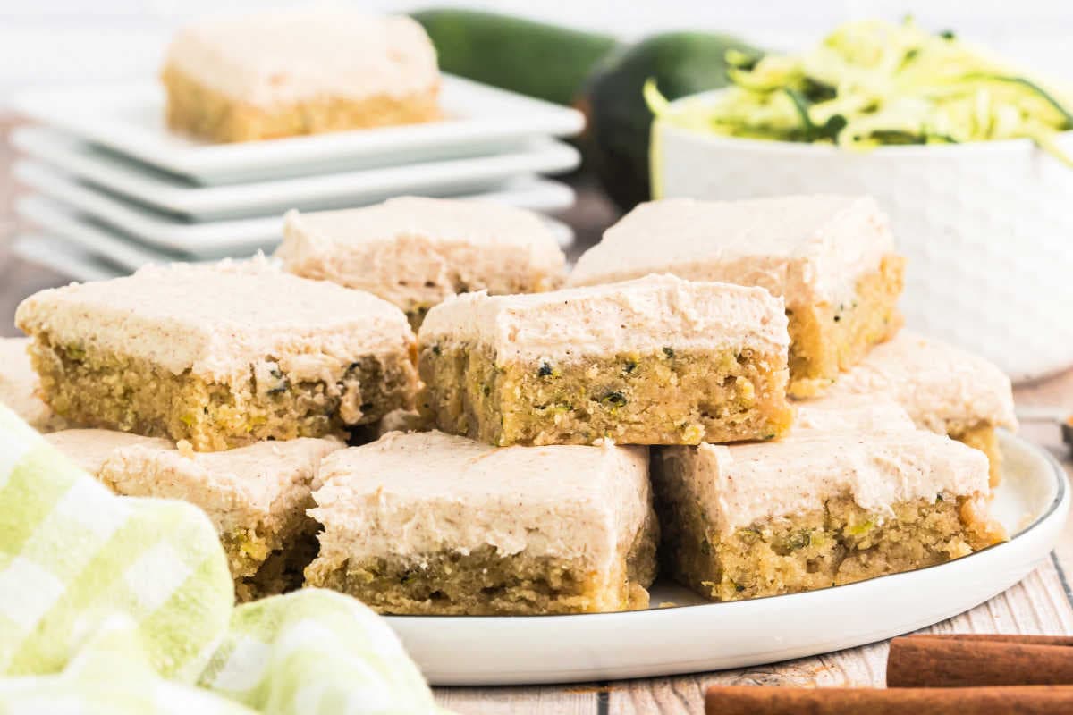 Cinnamon frosted zucchini cake bars stacked on a plate, with shredded zucchini and plates in the background.
