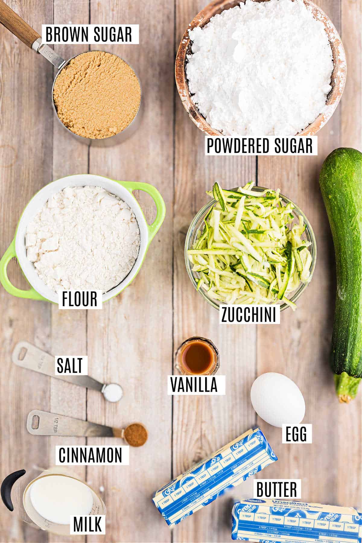 Baking ingredients on a wooden table, including sugars, flour, zucchini, butter, egg, milk, and spices—perfect for making cinnamon frosted zucchini cake bars.