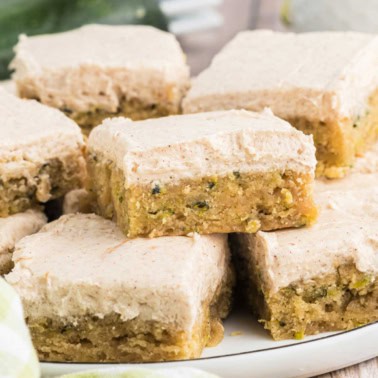 Cinnamon frosted zucchini cake bars are stacked on a white plate, revealing their moist, dense texture and luscious creamy frosting.