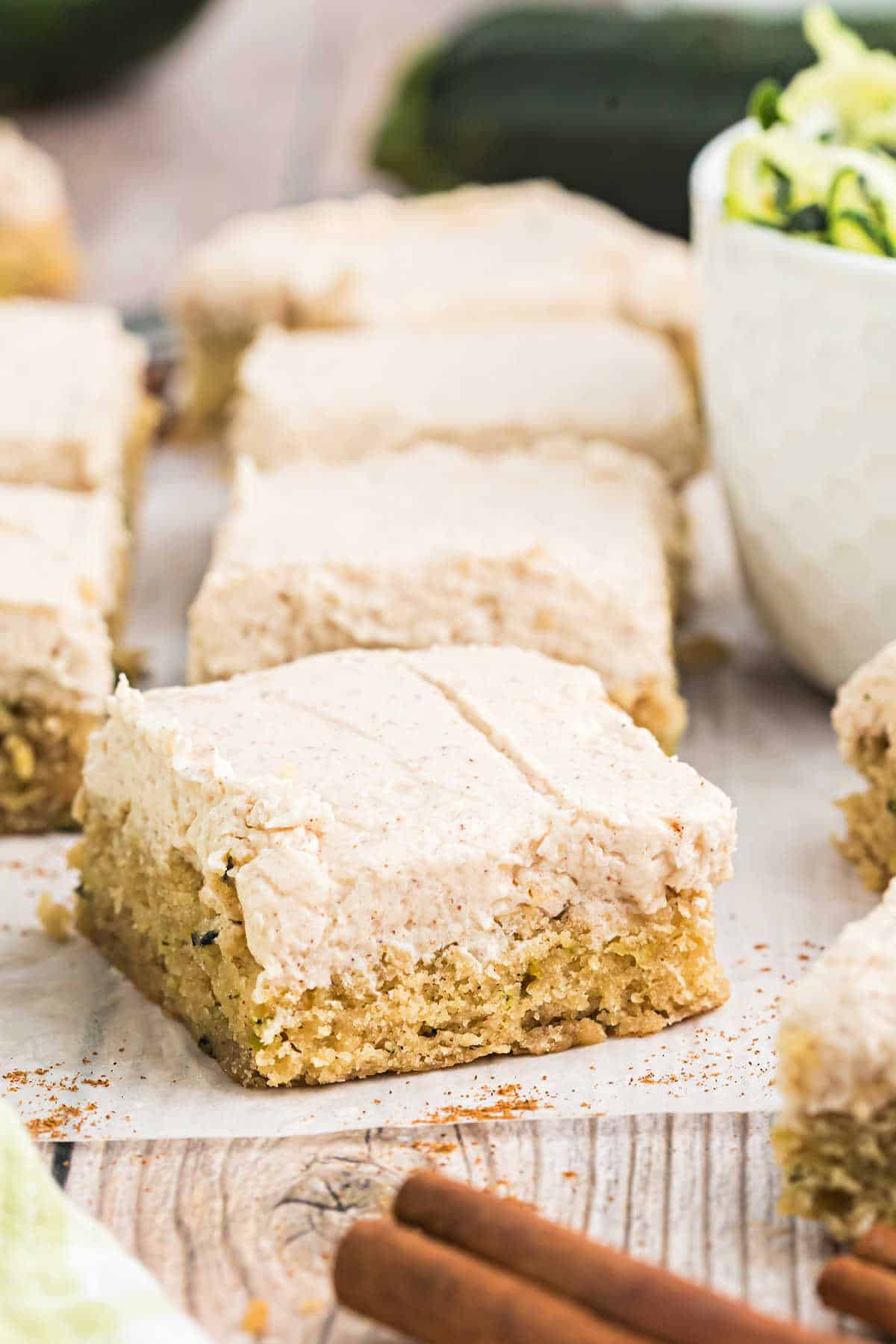 Cinnamon frosted zucchini cake bars rest on a wooden surface, with cinnamon sticks in the foreground.