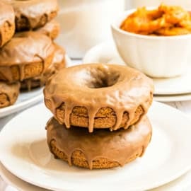 Two maple glazed pumpkin donuts stacked on a white plate, with more donuts and a bowl of orange filling in the background.