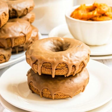 Two maple glazed pumpkin donuts stacked on a white plate, with more donuts and a bowl of orange filling in the background.
