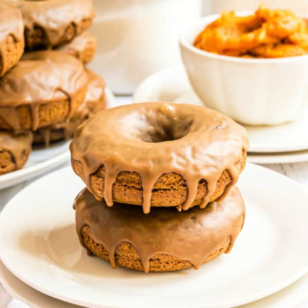 Two maple glazed pumpkin donuts stacked on a white plate, with more donuts and a bowl of orange filling in the background.