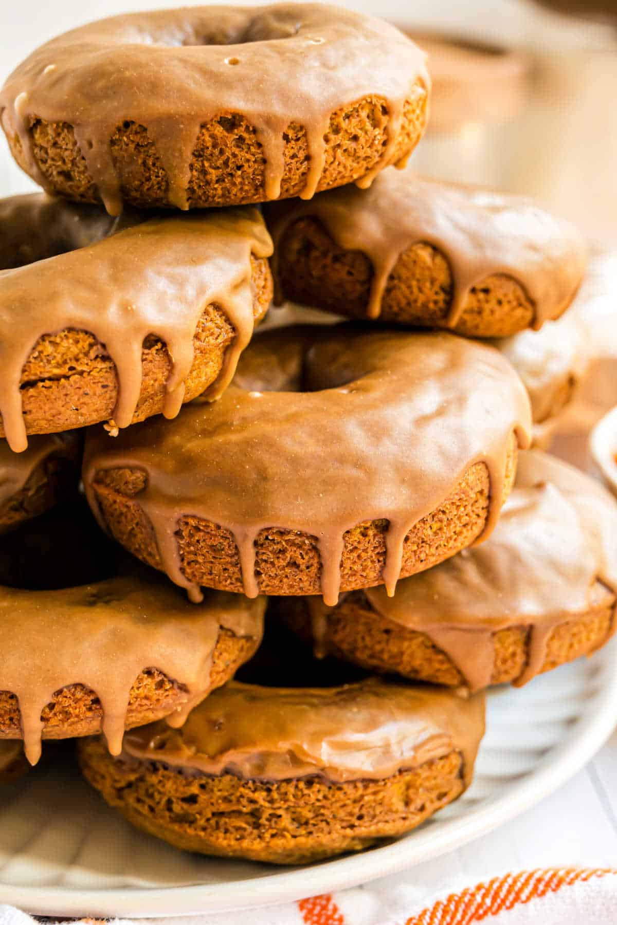 A stack of maple glazed pumpkin donuts with a shiny brown glaze dripping down the sides, on a white plate.
