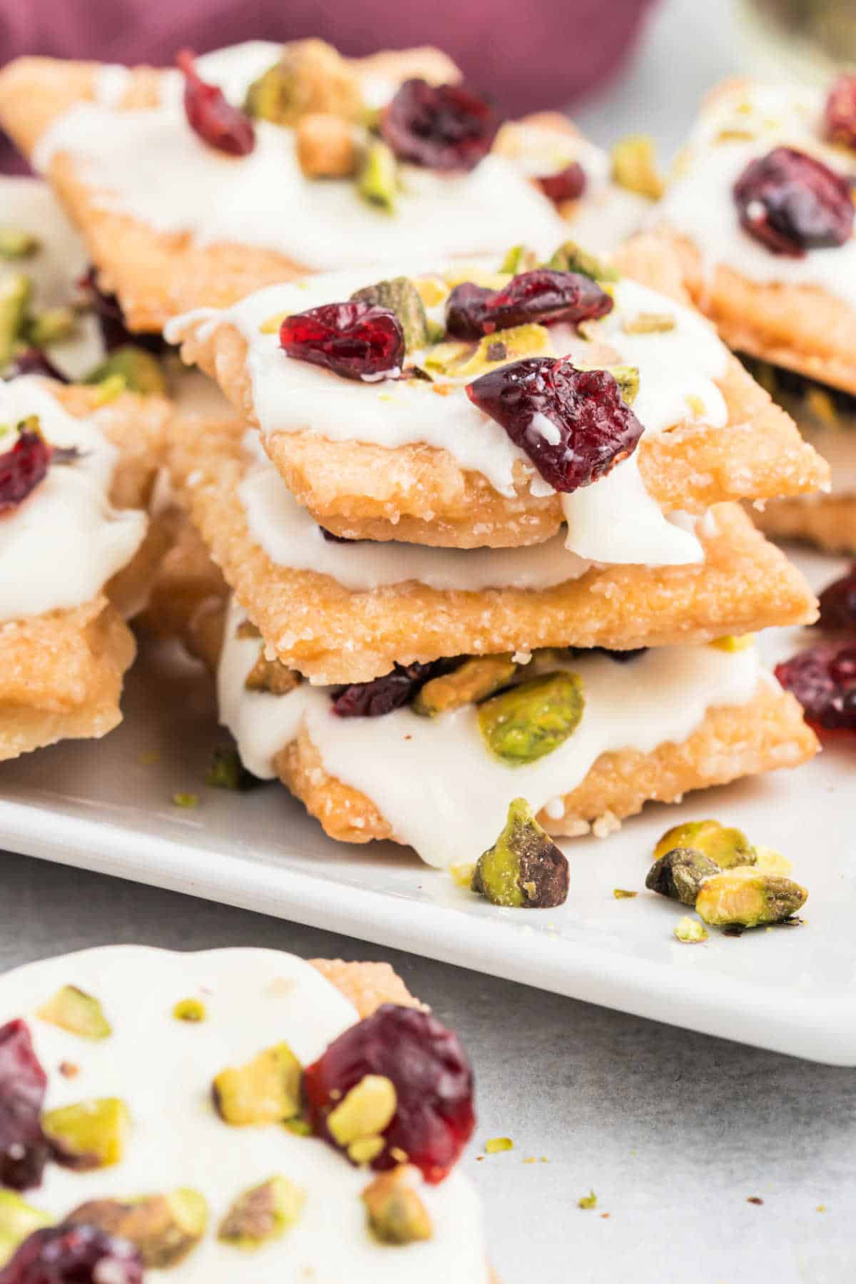 Stacked cranberry pistachio saltine toffee crackers coated with white icing, topped with dried cranberries and chopped pistachios on a white plate.