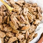 A gold scoop lifts Maple Brown Sugar Pecans from a bowl lined with white parchment paper.