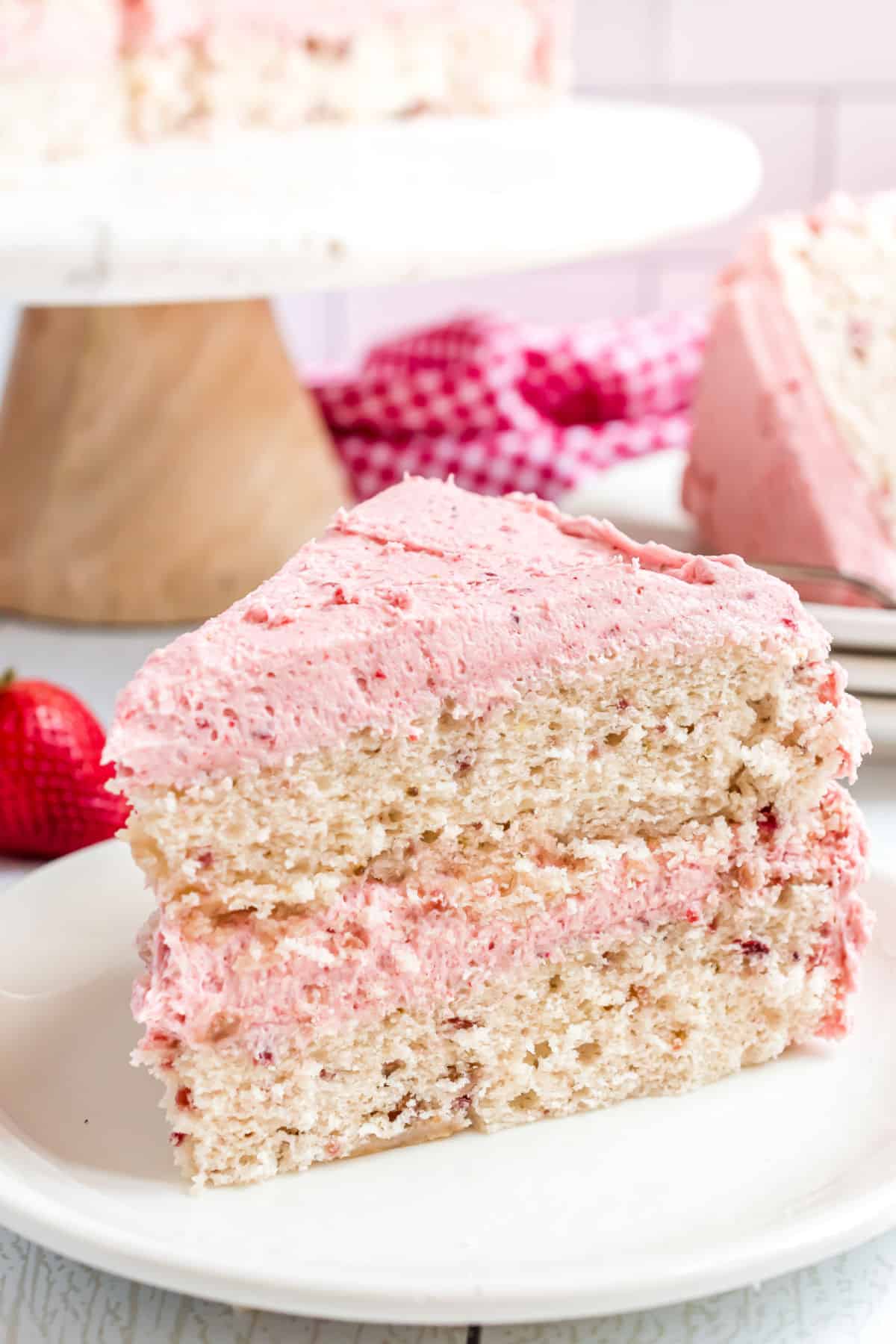 Slice of strawberry layer cake with strawberry frosting on white dessert plate with cake stand in background.