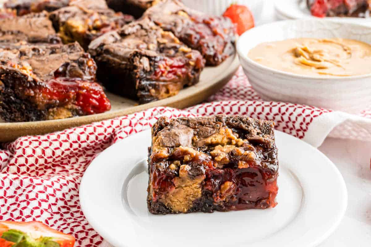 A brownie square with gooey cherry and peanut butter filling on a white plate, these irresistible peanut butter jelly brownies have more bars stacked in the background.