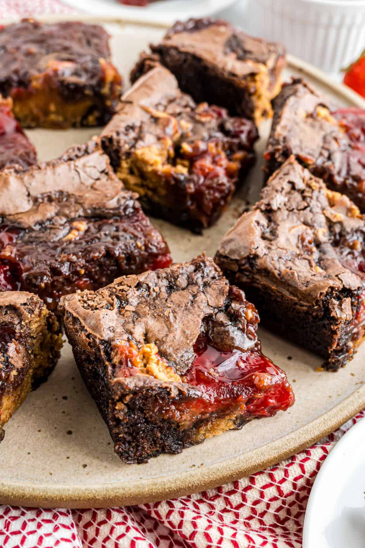 Peanut butter jelly brownies cut into squares, swirled with creamy peanut butter and bright red jelly, rest on a beige plate atop a red-patterned cloth.