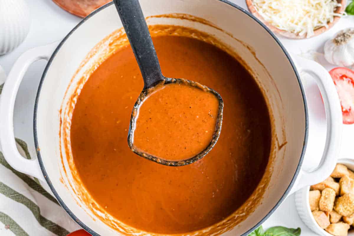 A ladle lifting creamy roasted tomato soup from a white pot, surrounded by croutons, cheese, and fresh ingredients.