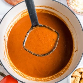 Ladle lifting creamy roasted tomato soup from a pot, with croutons and cheese on the side.