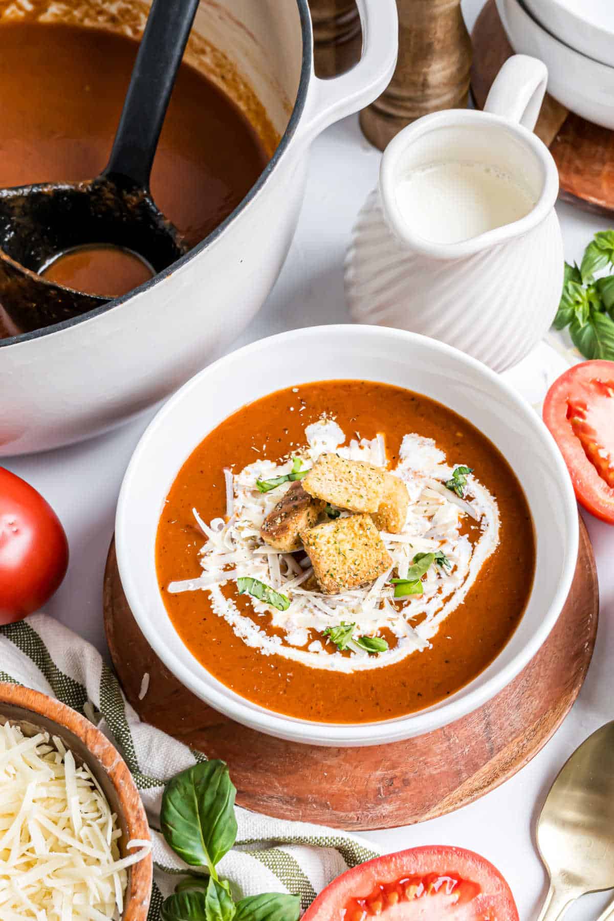 Bowl of roasted tomato soup with cream, croutons, and cheese, surrounded by fresh tomatoes, basil, and a pot of soup.