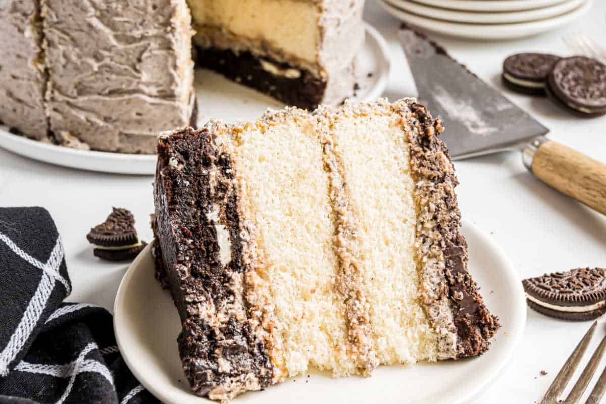 A slice of layered cookies and cream cake on a plate, with a whole oreo brownie cake and Oreos in the background.