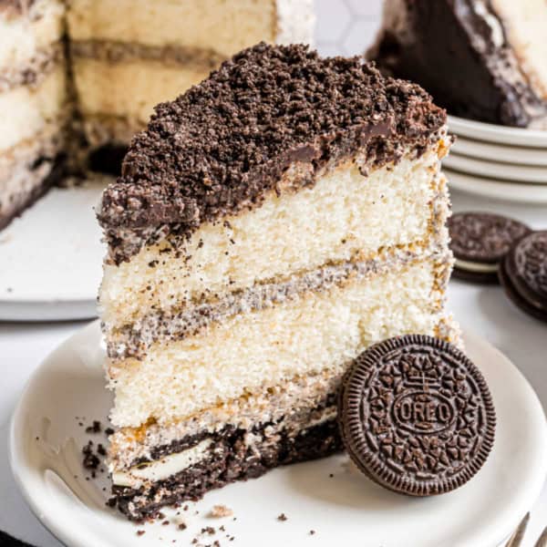 A slice of layered oreo brownie cake with cookies and cream, topped with Oreo cookie crumbs and a whole Oreo, sits on a white plate.