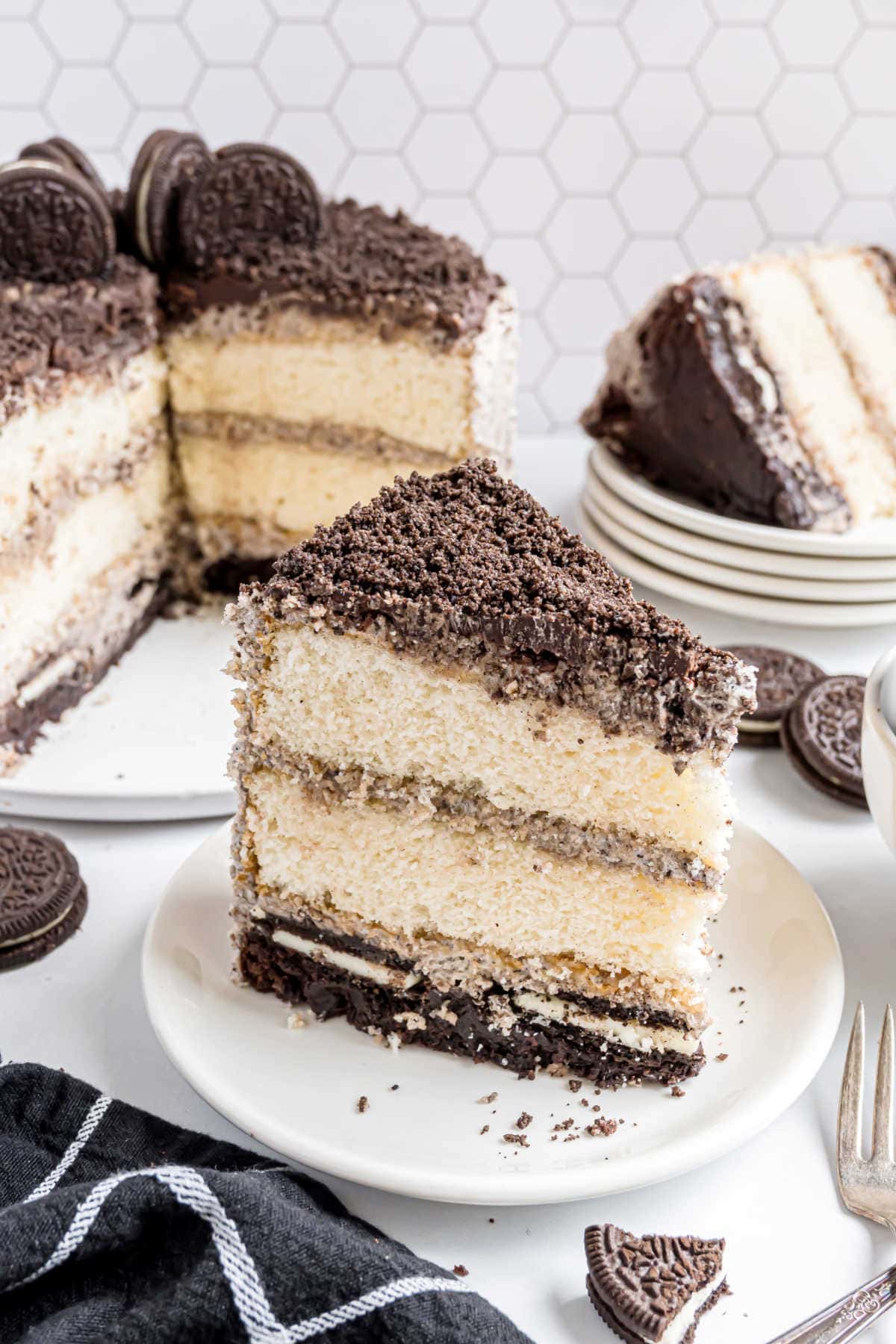 A slice of cookies and cream layer cake on a plate, with an Oreo brownie cake and cookies in the background.