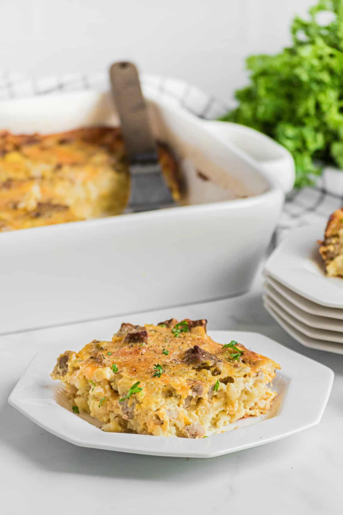 Slice of egg casserole on white plate, with dish of remaining casserole in background with fresh parsley.