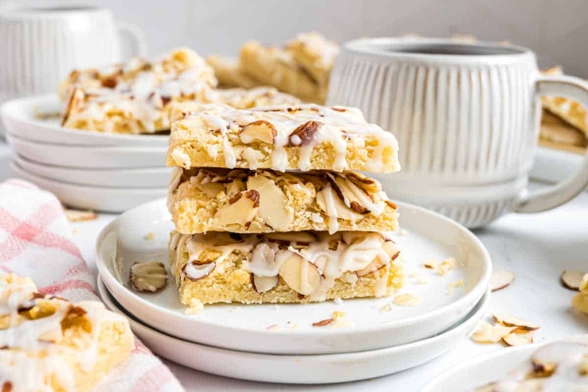 Three almond-topped dessert bars, made from a delicious almond bars recipe, are stacked on a plate next to a mug and scattered almond slices.