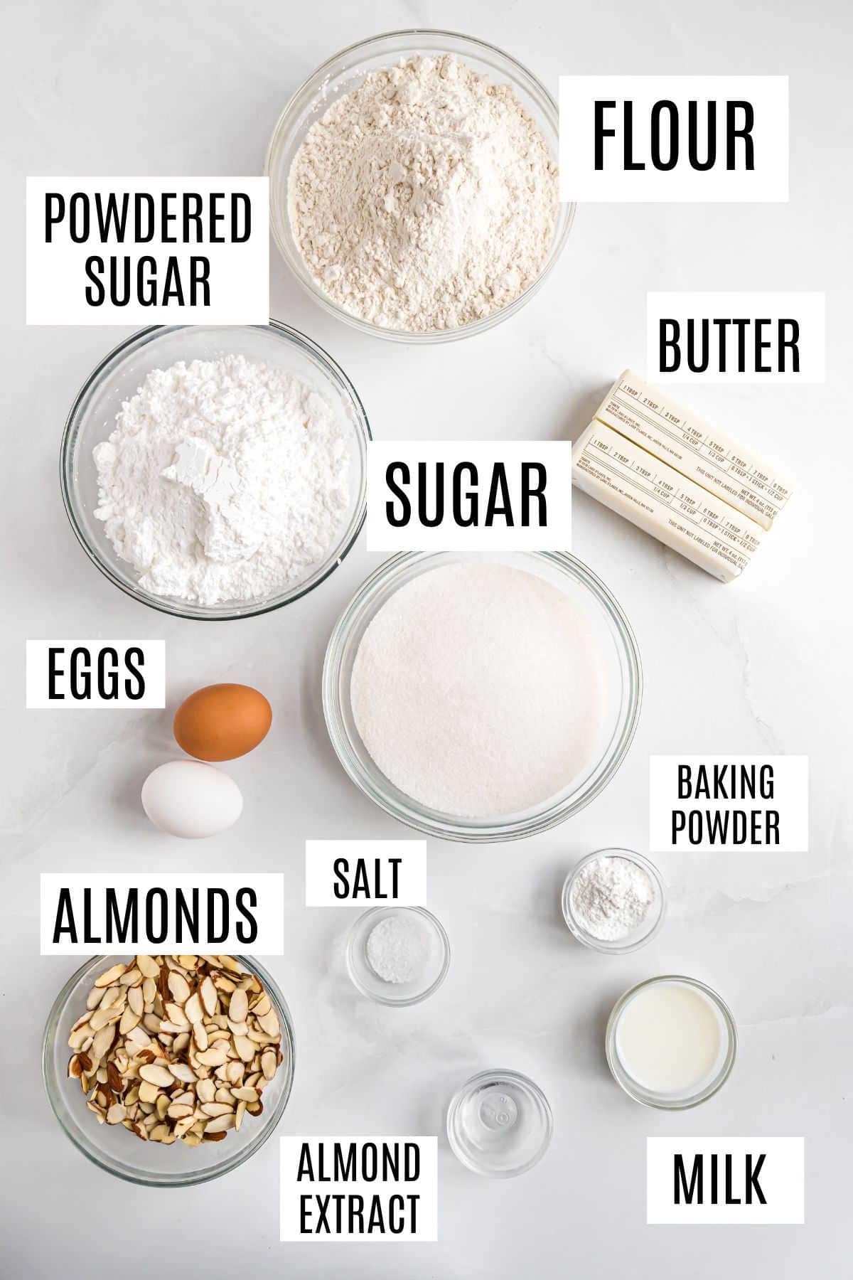 Bowls of flour, powdered sugar, sugar, almonds, baking powder, salt, butter, eggs, almond extract, and milk—everything you need for a classic almond bars recipe.