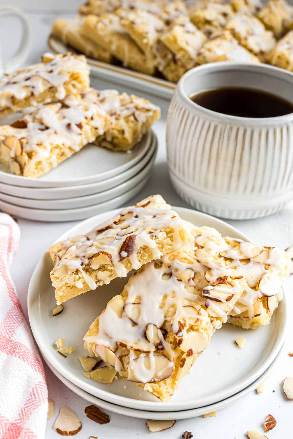 Two almond bars made from a delicious almond bars recipe, topped with white icing, rest on a plate beside a cup of black coffee and more pastries in the background.