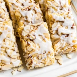 Rectangular almond pastry bars topped with sliced almonds and white icing, arranged on a white plate—an irresistible treat inspired by a classic almond bars recipe.