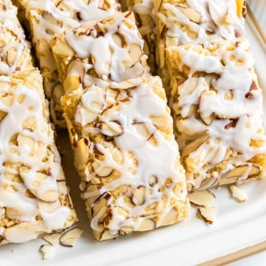 Rectangular almond pastry bars topped with sliced almonds and white icing, arranged on a white plate—an irresistible treat inspired by a classic almond bars recipe.